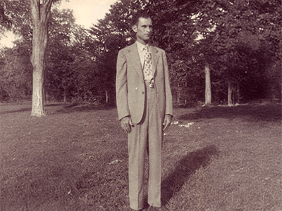 Black and white photo of a man in a light-colored suit and patterned tie standing on grass with trees in the background, conveying a formal and timeless tone.