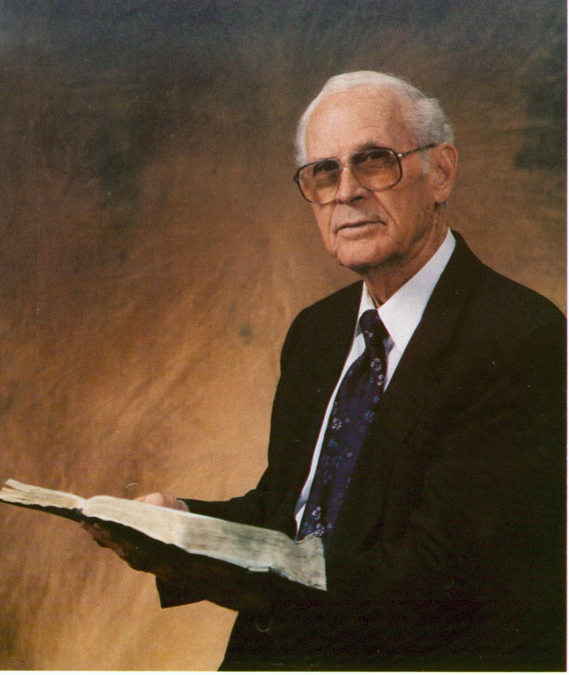 An elderly man in a suit and tie holds an open book, wearing glasses, with a thoughtful expression. The background is a gradient of warm tones.