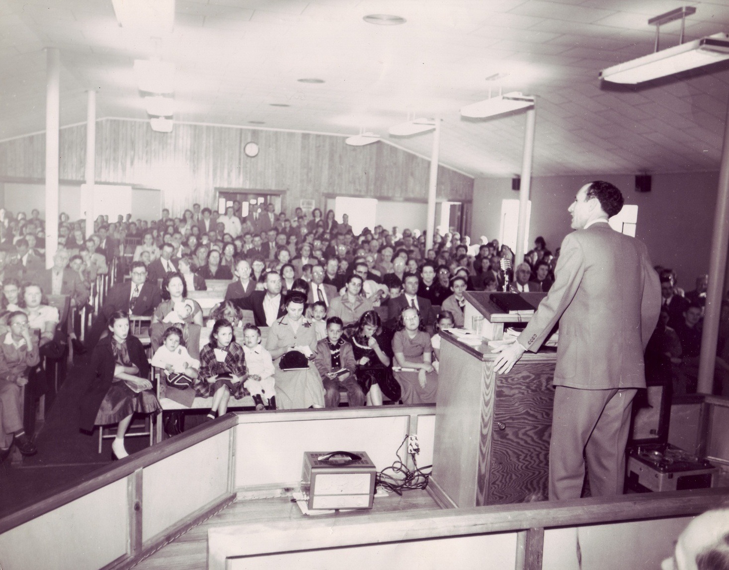 A man speaks at a podium in a packed church. The diverse congregation, dressed in 1950s attire, listens attentively. The atmosphere is engaged and respectful.