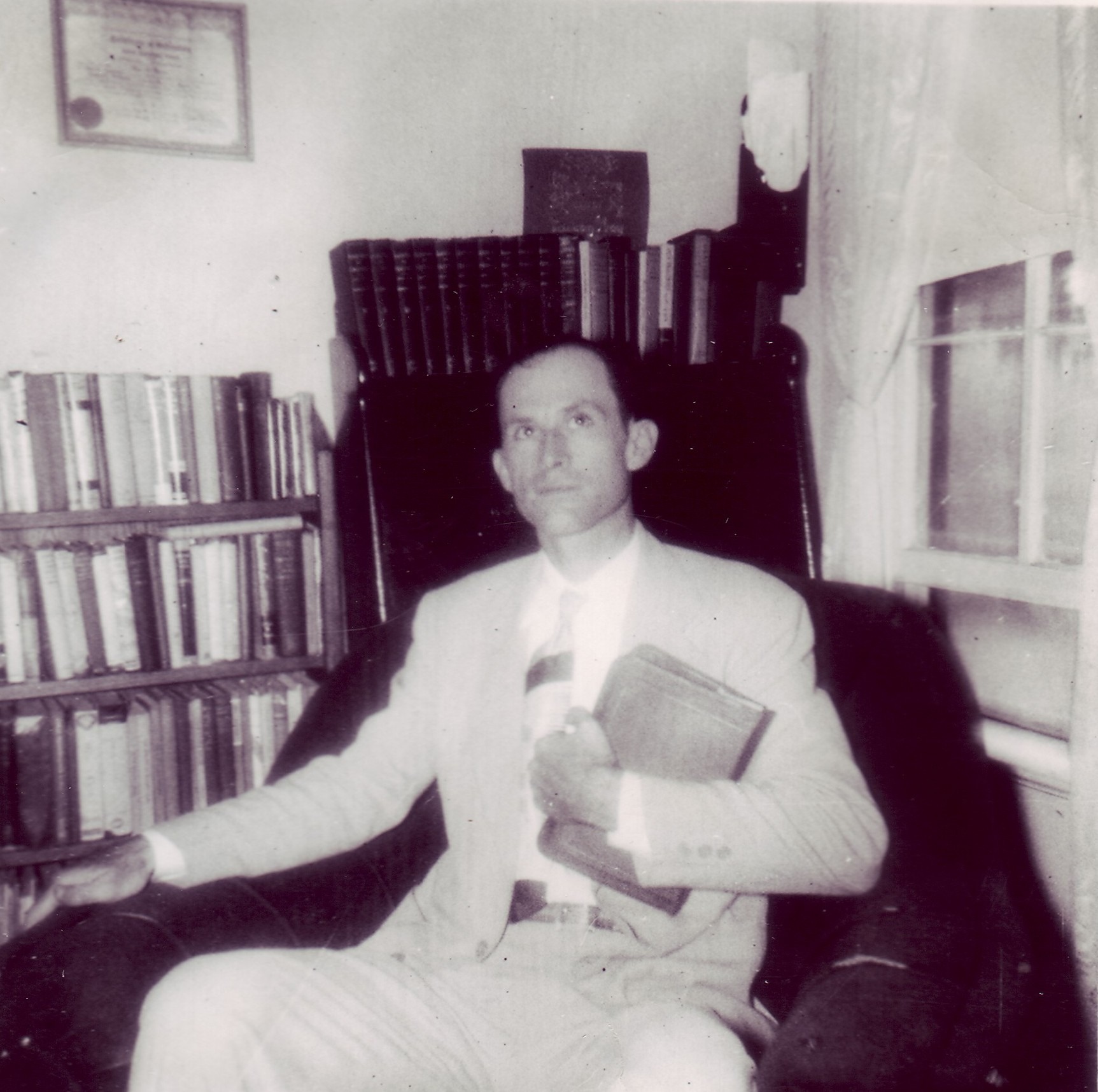 A man in a suit holds a book, seated in an armchair beside a bookshelf filled with books. The room exudes a scholarly, introspective atmosphere.