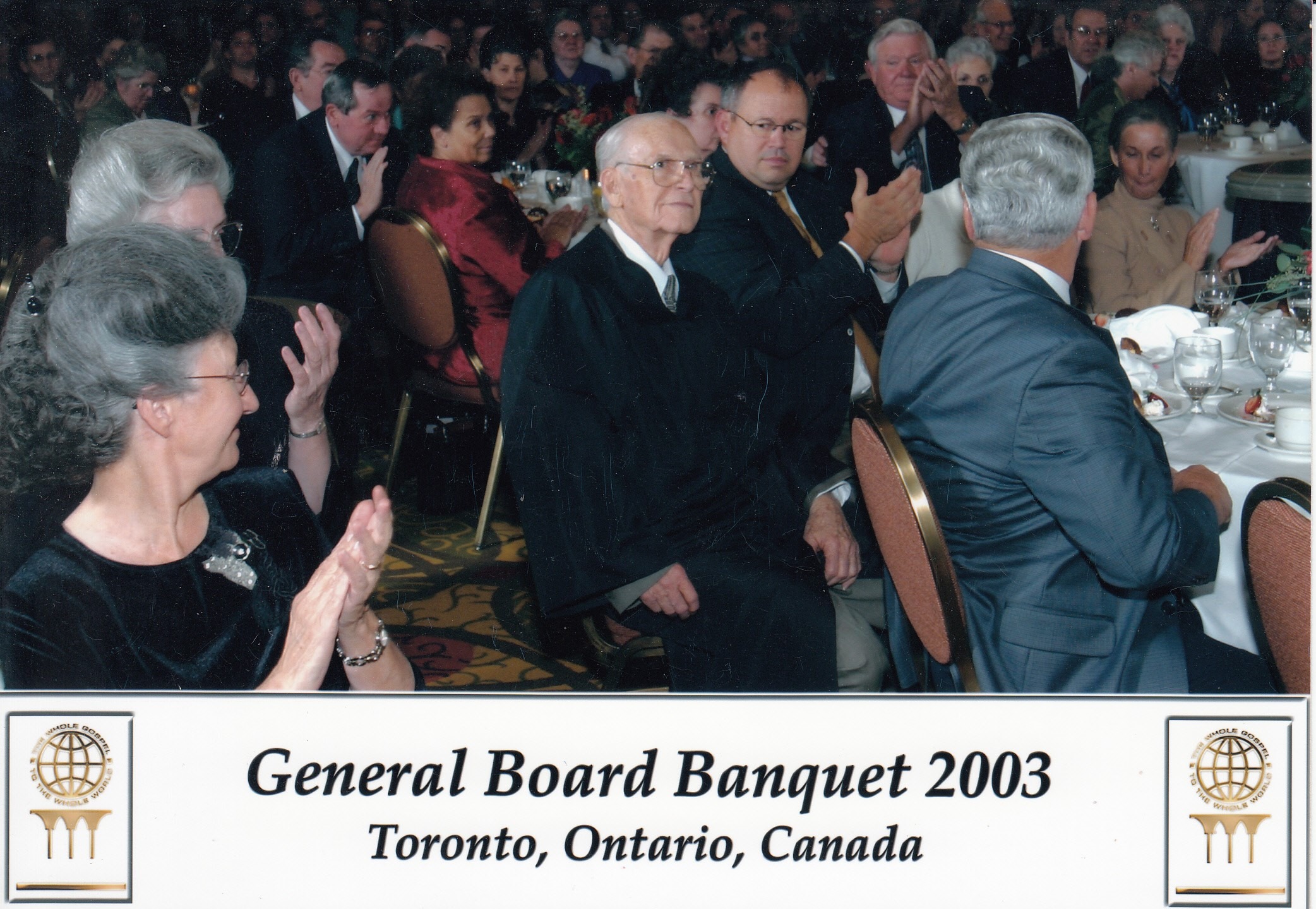 A formal banquet with attendees clapping. An elderly man in the center wears a black robe. The event is labeled "General Board Banquet 2003, Toronto, Ontario, Canada."
